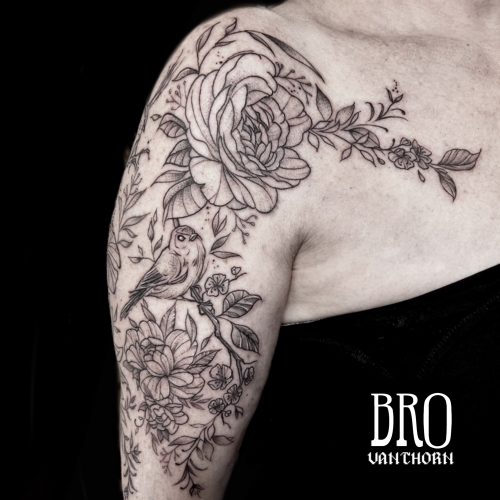 Sketch-style black and gray floral tattoo with blooming flowers and a small bird perched on a branch, covering the upper arm