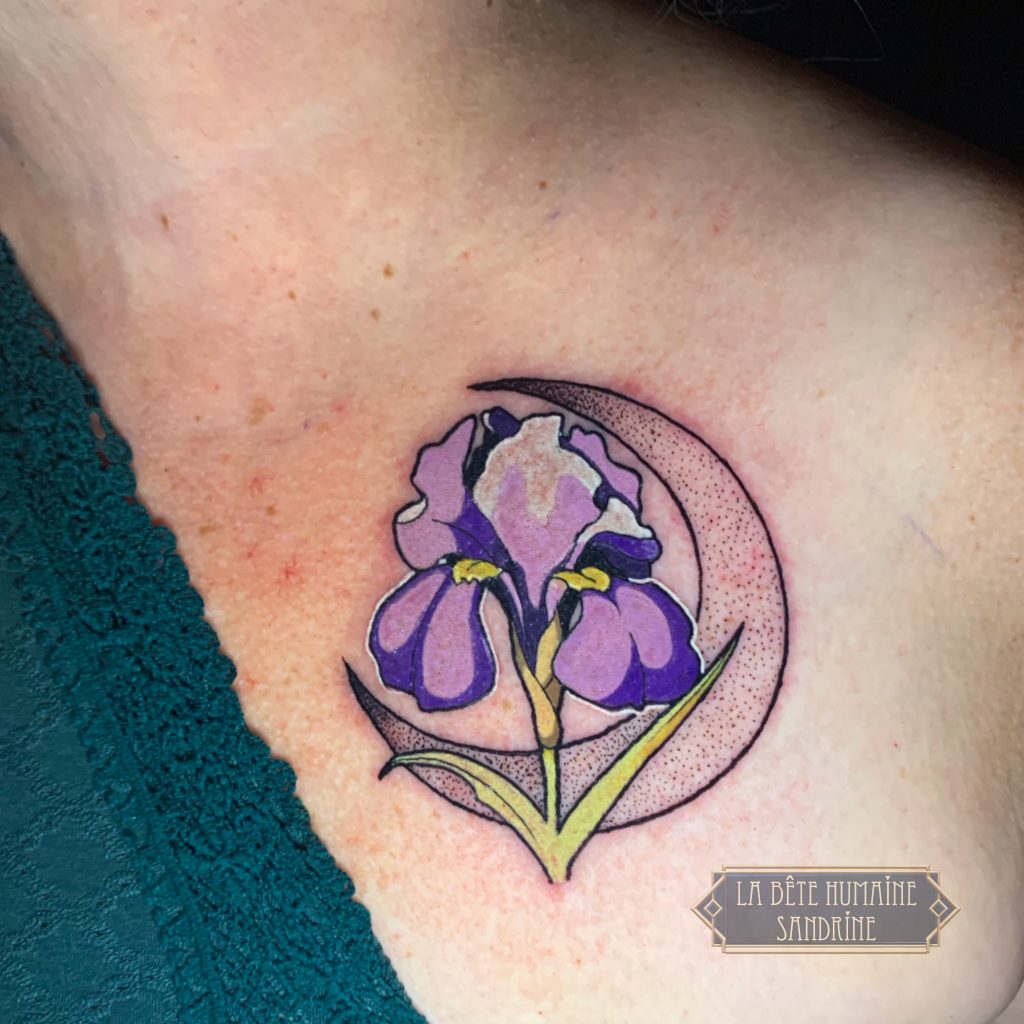 Tatouage d’un iris violet et vert, encadré par un croissant de lune en dotwork sur la poitrine, réalisé par Sandrine