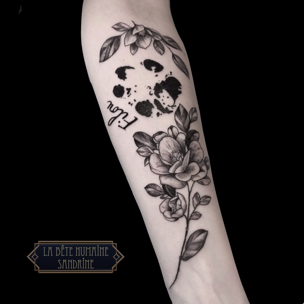 Avant-bras tatoué d’une élégante branche de fleurs en noir et gris encadrant une empreinte de patte stylisée, réalisé par Sandrine au salon La Bête Humaine à Paris.