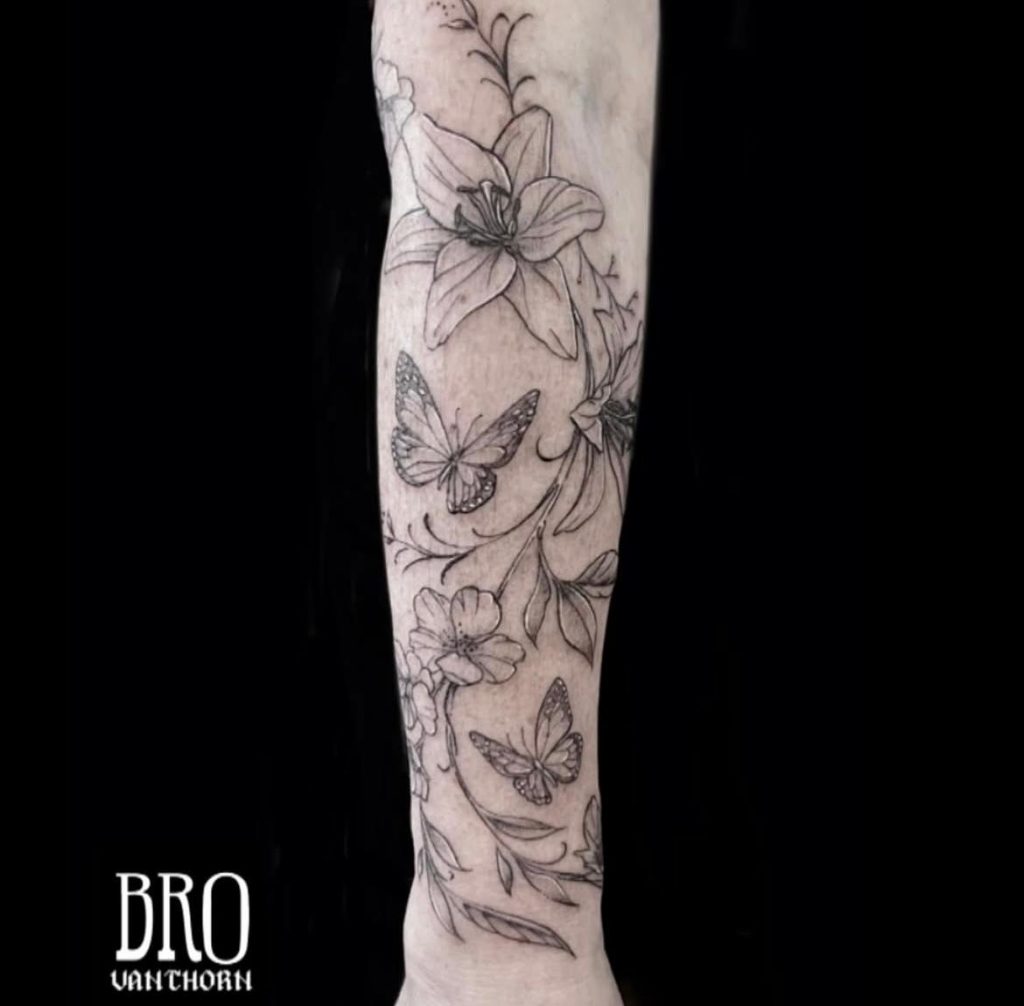Tatouages en noir et gris avec des lignes fines : grandes fleurs stylisées et trois papillons en vol, œuvre de Bro Vanthorn au salon La Bête Humaine.